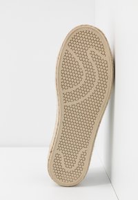 Semelle d'une chaussure beige avec un motif de grip circulaire, appuyée contre un coin blanc sur une surface lase.