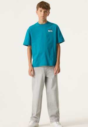 T-shirt turquoise à manches courtes avec le graphisme "DAYS", associé à un jean droit gris clair et des baskets blanches ; coupe et design décontractés.