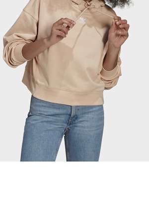 Personne portant un sweat à capuche beige Adidas et un jean bleu, ajustant les cordons de la capuche avec les deux mains.