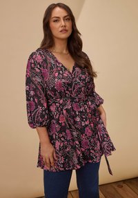 Donna con lunghi capelli castani che indossa una tunica nera con motivi floreali rosa e maniche a sbuffo, abbinata a jeans blu scuro, su uno sfondo beige.