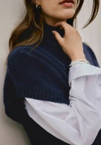 Eine Person trägt einen dunkelblauen Pullover über einem weißen Hemd, berührt ihren Hals, hat silberne Armbänder am Handgelenk und sichtbar gewelltes braunes Haar.