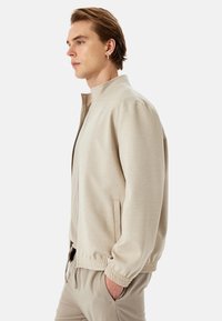 Chaqueta beige clara con cremallera, cuello alto, puños elásticos y bolsillos laterales. La tela tiene una textura suave y un corte relajado.