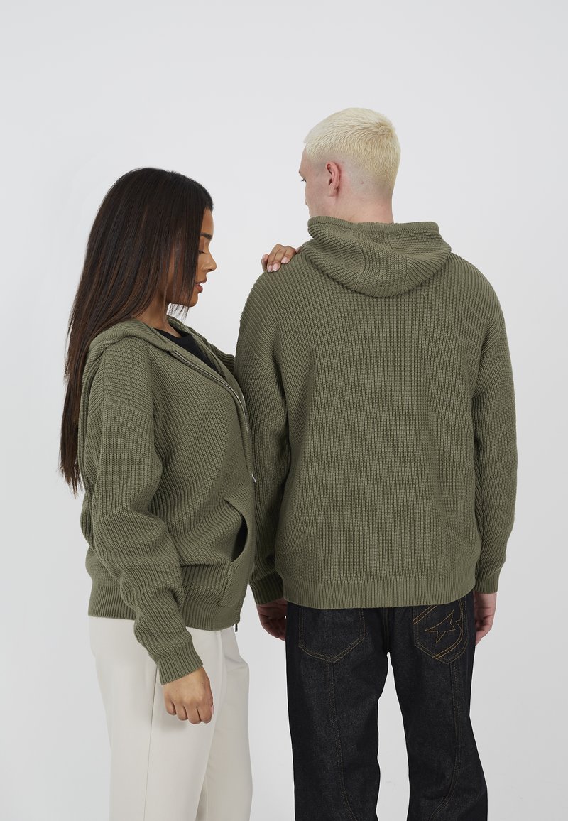 Olijfgroene geribbelde hoodie met ritssluiting en een relaxte pasvorm, met voorzakken, gedragen door twee personen, waarvan er één naar voren en de ander naar achteren kijkt.