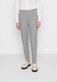Persoon in op maat gegoten grijze pantalon, witte blouse met lange mouwen en witte loafers met dikke zwarte zolen, staande tegen een eenvoudige achtergrond.