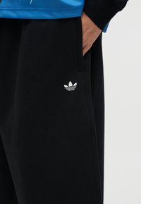 Osoba koja nosi crne hlače s bijelim Adidas logom na nozi i rukom u džepu, plavo-crni gornji dio djelomično vidljiv.