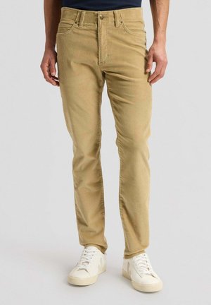 Mann trägt beige Cordhosen und weiße Turnschuhe und steht vor einem schlichten hellen Hintergrund.