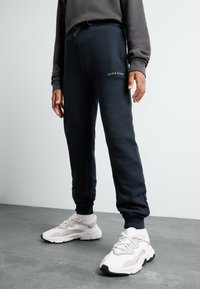 Pantalons de survêtement navy avec une coupe ajustée, taille élastique, poches latérales et détail de logo blanc. Associés à des chaussures de sport de couleur claire.