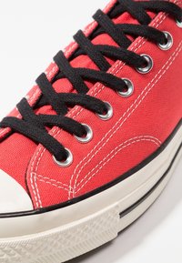 Baskets en toile rouge avec des lacets noirs, des œillets en métal argenté et un capuchon en caoutchouc blanc. Présente une semelle noire avec un grip texturé et des accents de couture blanche.