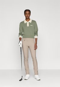 Zaļš adīts garo piedurknu polo ar krēmkrāsas apkakli un trim pogām, apvienots ar bēšiem šūtiem biksēm un baltām golfa kurpēm, tur rokā golfa nūju.