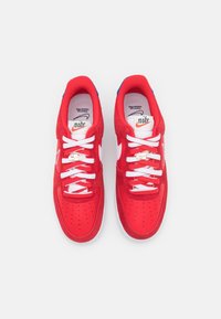 Červené semišové tenisky s bílými tkaničkami, perforovanou špičkou a po stranách, s bílým logem Nike a modrými akcenty na patách.