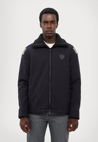 Svetlo modra zip-up jakna s mehkimi ovratnikom, oznako in črtastimi detajli na ramenih. Narejena je iz gladkega blaga in ima stranske žepe.