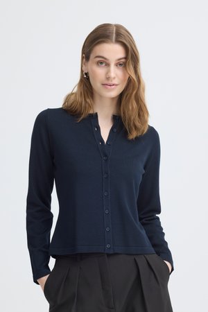 Õlgadeni helepruuni juustega naine, kes kannab tumesinist nööpidega kardigani ja musti volditud pükse, seisab käed taskutes.