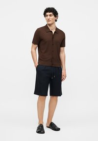 Mann steht mit den Händen in den Taschen, trägt ein braunes kurzärmeliges Hemd mit Knöpfen, marineblaue Shorts und schwarze Slip-on-Sandalen vor weißem Hintergrund.
