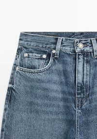 Blauwe denim jeans tailleband met lussen voor een riem, een zilverkleurige knoop, voorzak en muntzakje, met zichtbare textuur en stikdetails.