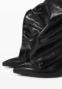 Bocanci negri din piele, cu un design căzut, vârful ascuțit și tocuri plate. Prezentare a nasturilor negri rotunzi aproape de gleznă.