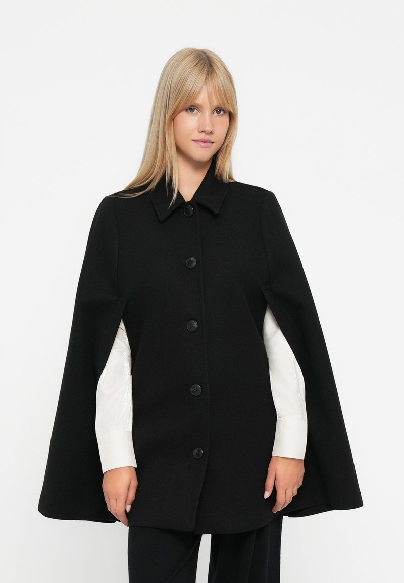 Cappotto di lana nera con colletto, sei grandi bottoni e fianchi aperti, indossato sopra una camicia bianca. Texture liscia e design minimale.