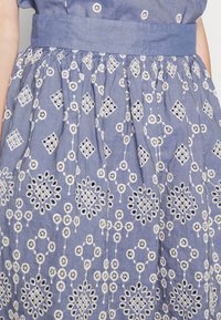 Modrá chambray šaty s bílými vyšívanými květinovými vzory a geometrickými akcenty, se skládací pasem a plochým pasem.