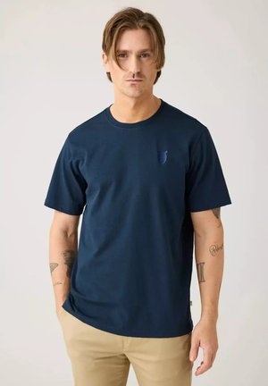 Hombre con cabello de longitud media que lleva una camiseta azul marino y pantalones beige, de pie contra un fondo claro y liso.