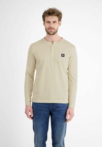 Beige Langarmhemd mit Henley-Ausschnitt, versehen mit einem kleinen schwarzen Logo-Patch auf der linken Brust. Kombiniert mit blauen Jeans.