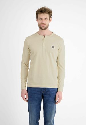 Beige longsleeve shirt met een henley-halslijn, voorzien van een klein zwart logopatch op de linkerborst. Gepaard met blauwe denim jeans.