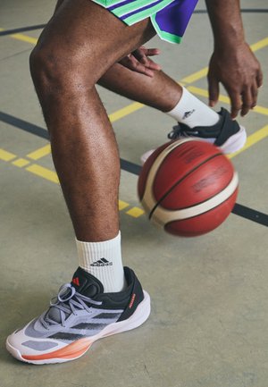 Chaussure de basketball avec tige grise, accents noirs et semelle orange. Chaussettes blanches avec logo Adidas noir. Ballon de basketball roulant sur un terrain.