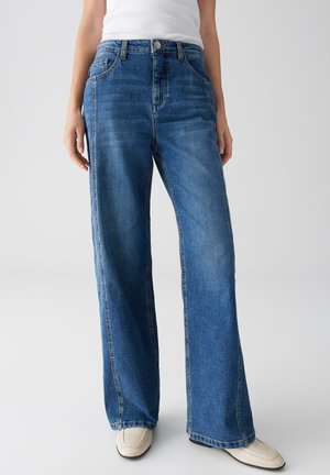 Jeans bleus à jambe large en denim, dotés d'une fermeture à bouton et à glissière, de cinq poches et de coutures visibles le long des coutures.