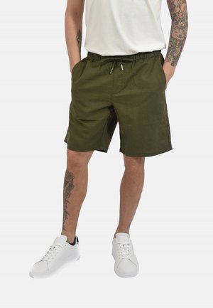 Uomo che indossa pantaloncini verde oliva con coulisse, scarpe da ginnastica bianche, maglietta bianca e tatuaggi su braccia e gambe, in piedi con le mani in tasca.