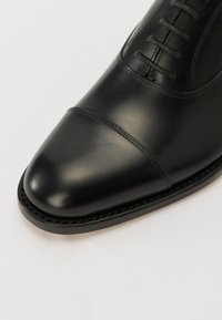 Zapato de cuero negro con punta redondeada, que presenta cordones y un diseño de puntera cosida. La suela tiene un borde suave.