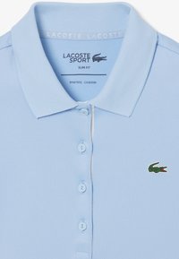 Polo de color azul claro, confeccionado en una tela suave y de corte ajustado. Presenta un collar de canalé, tapeta con cinco botones y un pequeño logo de cocodrilo verde.