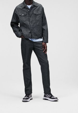Homme portant une veste et un pantalon assortis noirs et gris à motifs avec des baskets noires et blanches, debout devant un fond uni.