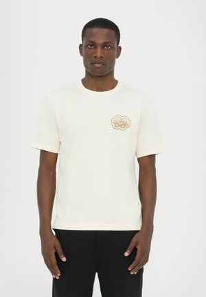 Cremefarvet bomulds t-shirt med rund halsudskæring og korte ærmer, der har et tekstureret beige logo på venstre bryst.
