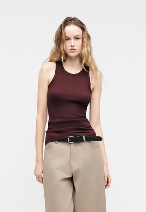 Jeune femme aux longs cheveux ondulés portant un haut bordeaux sans manches, un pantalon beige et une ceinture noire sur un fond clair uni.