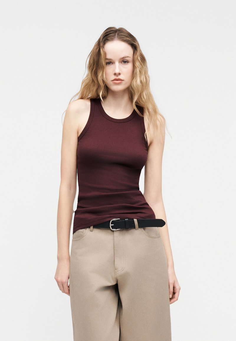 Jeune femme aux longs cheveux ondulés portant un haut bordeaux sans manches, un pantalon beige et une ceinture noire sur un fond clair uni.