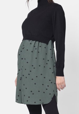 Kvinde iført en sort cropped sweater over en grøn knappet kjole med sorte polkaprikker, kombineret med sorte leggings.
