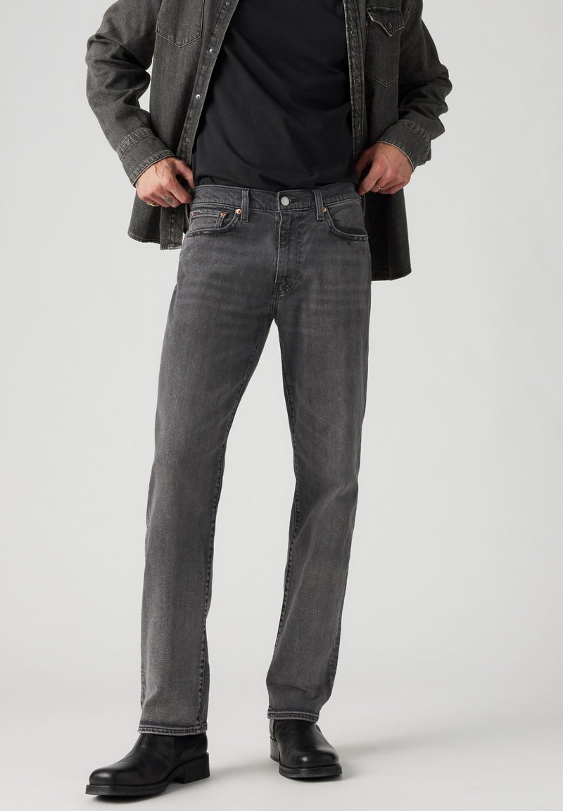 Uomo in jeans grigi sbiaditi, t-shirt nera, giacca di jeans scura e stivali di pelle neri in piedi con le mani vicino alla vita.