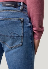 Blaue Jeans aus Denim verfügen über eine Gesäßtasche mit einem schwarzen Schriftlogo. Texturiertes Material mit leichter Abnutzung und schmaler Schnitt ab der Taille.
