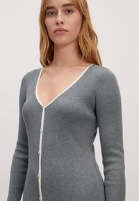 Grauer gerippten Cardigan mit tiefem V-Ausschnitt und cremefarbigen Knopfakzenten. Lange Ärmel, taillierte Form und weiche Textur.