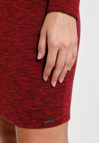 Robe en tricot rouge présentant un motif texturé, une coupe ajustée et une petite étiquette de marque près de l'ourlet. Gros plan de la robe et de la main.