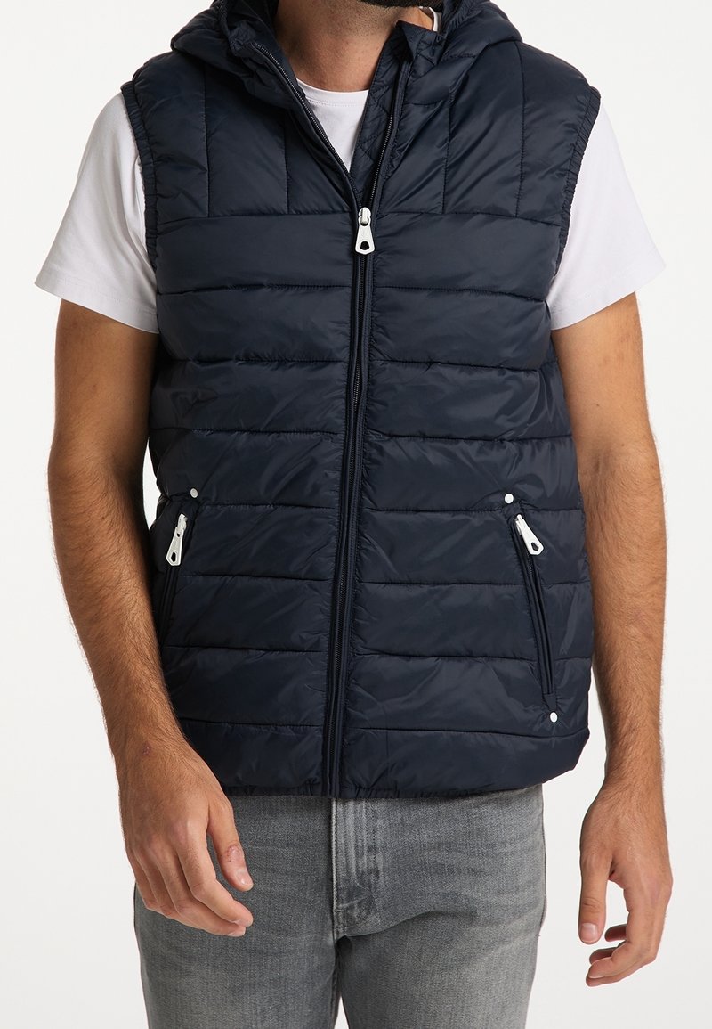 Homme portant un gilet noir sans manches à capuche, matelassé, sur un t-shirt blanc à manches courtes et un jean gris, debout avec les bras détendus.