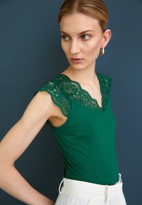 Green tank top with lace detail at the shoulder and neckline, featuring a fitted design and soft fabric. Paired with light-colored trousers.