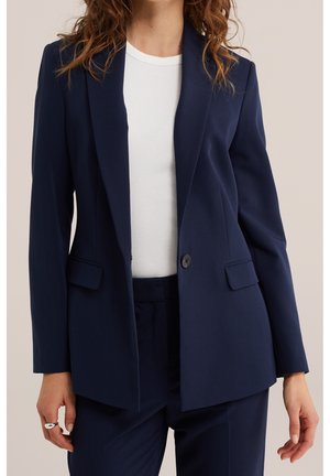 Vrouw draagt een marineblauwe blazer met één knoop dichtgespeld over een wit geribd topje, gecombineerd met bijpassende marineblauwe broek.