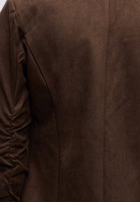 Gros plan d’un tissu en daim marron avec un détail de couture froncée sur le côté gauche, montrant la texture et les lignes de couture.