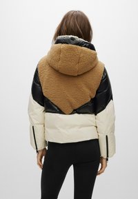 Giacca imbottita nera, crema e marrone con cappuccio soffice e strutturato. Presenta un design geometrico a blocchi di colore e tasche laterali con zip.