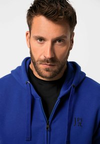 Sudadera azul con cremallera y cordones, cremallera negra y un logo en el pecho. Presenta una textura suave y un diseño casual.