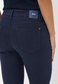 Tmavě modré džíny s hladkým povrchem. Mezi jejich vlastnosti patří zadní kapsy, štítek na pasu s logem a decentní švy.