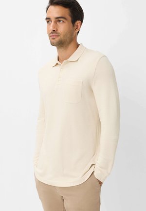 Man draagt een crèmekleurig poloshirt met lange mouwen en een borstzakje, beige broek, staand met handen in de zakken tegen een witte achtergrond.