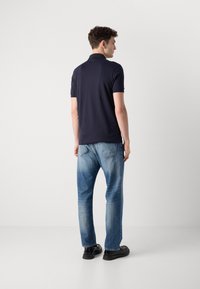 Jūrų mėlynumo polo marškiniai su trumpomis rankovėmis. Dėvimi su labai šviesiais mėlynais džinsais, užbaigti juodais batais. Atgalinis vaizdas prieš paprastą foną.