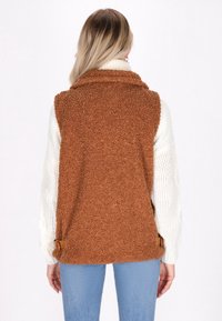 Braune, flauschige Weste mit Kragen, strukturiertem Material, seitlichen Akzenten und einem weißen, gestrickten Langarmshirt darunter. Rückansicht gezeigt.