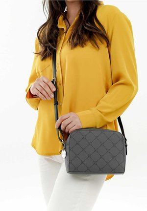 Kvinde iført en gul bluse og hvide bukser holder en lille grå crossbody taske med mønstret design og rund metalnøglebrik.