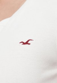 Λευκό ριμπ ύφασμα με μικρό, κεντημένο κόκκινο λογότυπο γλάρου στη αριστερή πλευρά ενός τοπ με V-λαιμόκοψη.